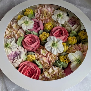 May include: A round, white framed floral arrangement. The arrangement features a variety of buttercream flowers in shades of pink, yellow, white, and green. The flowers are arranged in a dense, textured pattern, creating a visually appealing display.