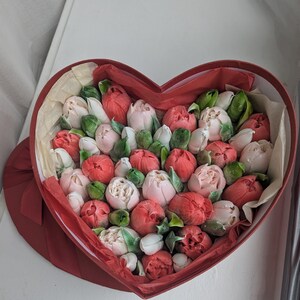 May include: A heart-shaped red box filled with a collection of small, rounded flower buds in shades of red, pink, and white. The buds are interspersed with small green leaves and nestled within light-colored tissue paper inside the container.