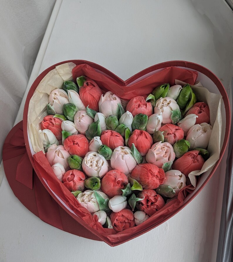 May include: A heart-shaped red box filled with a collection of small, rounded flower buds in shades of red, pink, and white. The buds are interspersed with small green leaves and nestled within light-colored tissue paper inside the container.