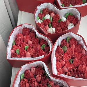 May include: Five heart-shaped red boxes filled with red and white tulip-shaped confections and green leaves. Each box is lined with white tissue paper, containing multiple edible flower-shaped treats arranged closely together in a dense cluster.