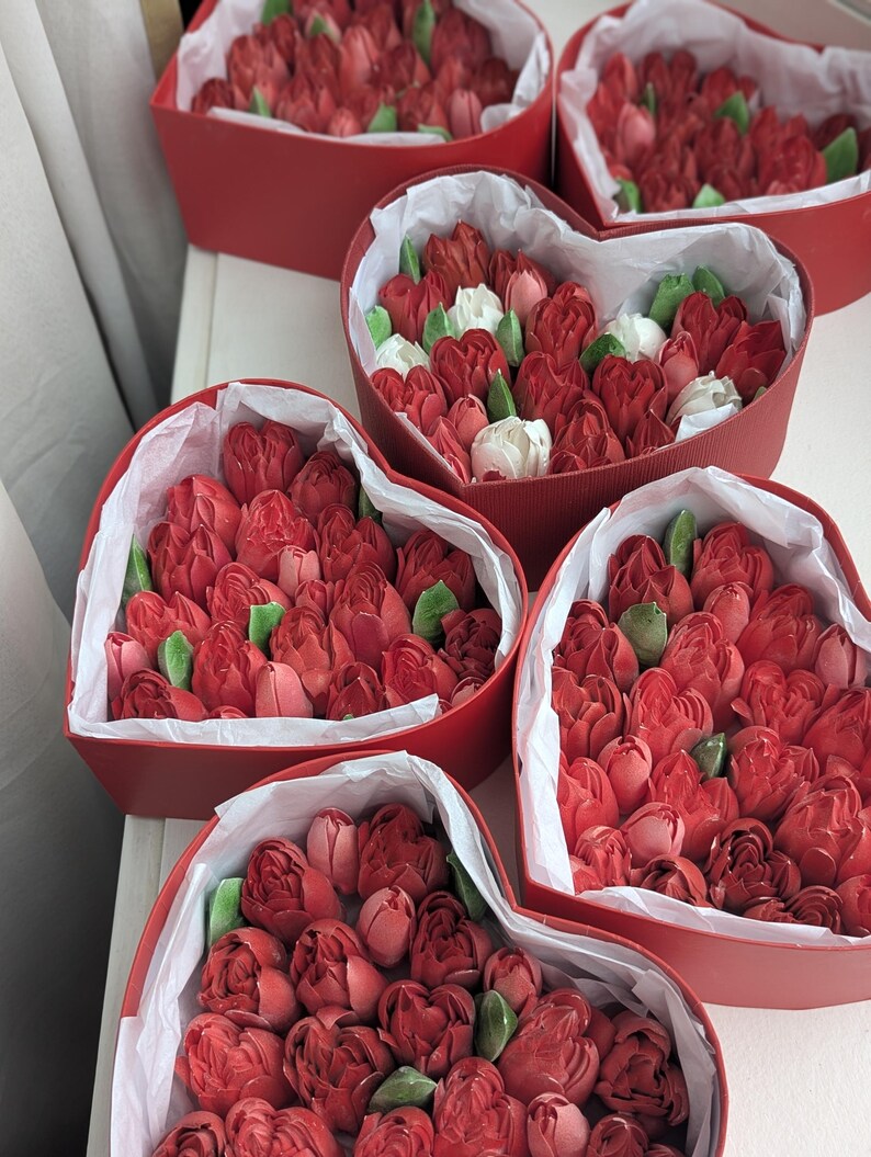 May include: Five heart-shaped red boxes filled with red and white tulip-shaped confections and green leaves. Each box is lined with white tissue paper, containing multiple edible flower-shaped treats arranged closely together in a dense cluster.