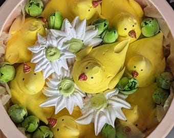 May include: A round, light pink gift box filled with yellow meringue chicks, white meringue flowers with green centres, and small green meringue buds. The chicks have small, pointed, reddish-brown beaks and dark eyes, arranged neatly inside the container.