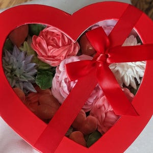 May include: A heart-shaped red gift box with a clear window lid, tied with a red satin ribbon bow. Inside are various floral-shaped confections in shades of pink, white, and lavender, along with small red heart-shaped pieces and green leaves.