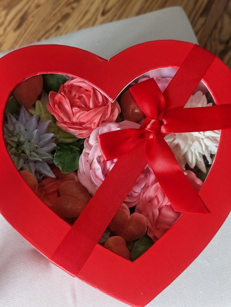 May include: A heart-shaped red gift box with a clear window lid, tied with a red satin ribbon bow. Inside are various floral-shaped confections in shades of pink, white, and lavender, along with small red heart-shaped pieces and green leaves.