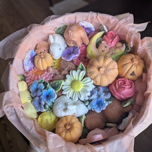 May include: A round container lined with light pink tissue paper, filled with an assortment of small, molded figures including orange pumpkins, white and brown rabbits, teddy bears, and various colorful flowers like daisies and roses.