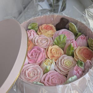 May include: A round, light pink box filled with edible roses in shades of pink, yellow, and peach. Green leaf-shaped candies are interspersed among the flowers. The box is partially open, revealing the contents.