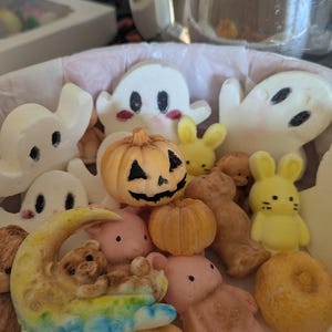 May include: A collection of small, molded decorative figures including white ghosts with black eyes, orange pumpkins with carved faces, yellow bunnies, pink bears, and a crescent moon with a bear. The items are arranged together in a container.