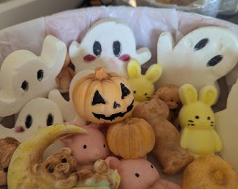 May include: A collection of small, moulded decorative figures including white ghosts with black eyes, orange pumpkins with carved faces, yellow bunnies, pink bears, and a crescent moon with a bear. The items are arranged together in a container.