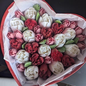 May include: A heart-shaped red box filled with a dense arrangement of white, deep red, and dusty pink flower-shaped confections. The treats are nestled in white tissue paper, featuring intricate petal details and small green leaf accents.