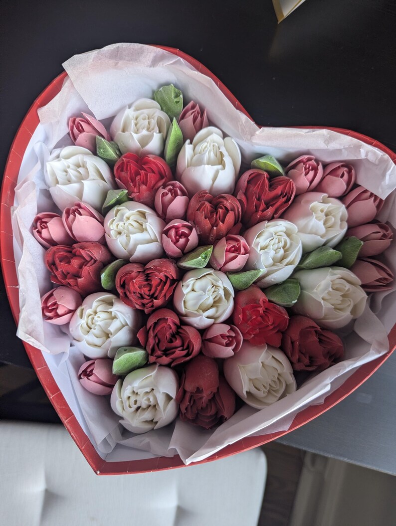 May include: A heart-shaped red box filled with a dense arrangement of white, deep red, and dusty pink flower-shaped confections. The treats are nestled in white tissue paper, featuring intricate petal details and small green leaf accents.