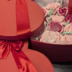 May include: Two red heart-shaped gift boxes with textured lids and shiny red satin ribbons. One box is open, revealing a collection of light pink and deep magenta rose-shaped confections accented with small, pale green leaf-shaped decorations.