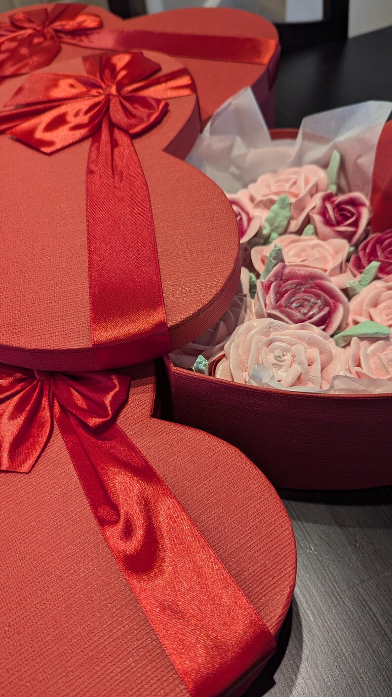 May include: Two red heart-shaped gift boxes with textured lids and shiny red satin ribbons. One box is open, revealing a collection of light pink and deep magenta rose-shaped confections accented with small, pale green leaf-shaped decorations.