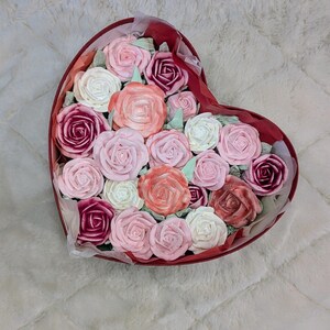 May include: A heart-shaped red box filled with an arrangement of rose-shaped items in various shades of pink, white, peach, and deep magenta. The roses are tightly packed together, creating a dense floral display within the container.