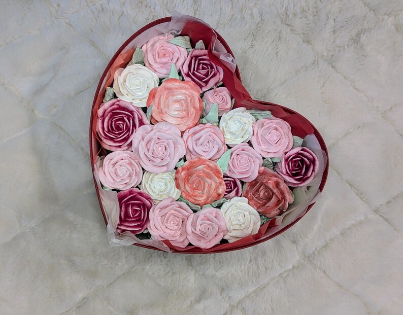 May include: A heart-shaped red box filled with an arrangement of rose-shaped items in various shades of pink, white, peach, and deep magenta. The roses are tightly packed together, creating a dense floral display within the container.
