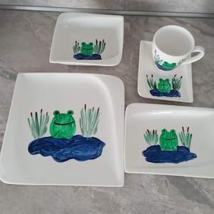 Könnte beinhalten: Ein weißes Keramik-Geschirrset mit Frosch-Design. Das Set umfasst eine quadratische Schüssel, eine Tasse mit Untertasse und drei Teller. Jedes Teil ist mit einem grünen Frosch auf einem blauen Teich mit grünen Schilf dekoriert.