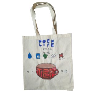 May include: Cream canvas tote bag with a hand-painted design. The design includes the text "WHEN LIFE GIVES YOU..." and "MAKE TEA". The illustration features a tea cup, tea leaves, and other tea-related icons.