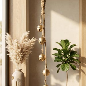 Op de afbeelding: Een hangende decoratie met vijf bolvormige, messingkleurige bellen bevestigd aan een gevlochten touw van jute. Het touw is aan de bovenkant geknoopt om op te hangen, waarbij de bellen verticaal langs het koord zijn verdeeld met elk een gleuf.