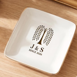Op de afbeelding: Een vierkant, wit keramisch sieradenschaaltje met gouden rand. Het schaaltje heeft een zwart wilgenboomontwerp met de initialen "J & S" en het jaar "SINCE 2026". Het schaaltje staat op een houten oppervlak.
