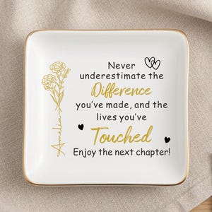Könnte beinhalten: Eine weiße Keramik-Ringschale mit goldenem Rand, mit einem Blumendesign und dem Text "Never underestimate the Difference you've made, and the lives you've Touched." Ein goldener Ring und eine Karte sind ebenfalls vorhanden.
