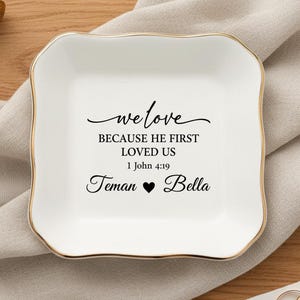May include: A white ceramic trinket dish with a gold rim and a square shape. The dish features the text "we love BECAUSE HE FIRST LOVED US 1 John 4:19 Teman ❤ Bella" in black script. The dish is on a wooden surface.