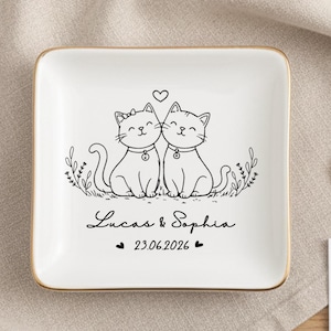 May include: A white ceramic ring dish with a gold rim, featuring a black line drawing of two cats with a heart above them. The dish is inscribed with the names "Lucas & Sophia" and the date "23.06.2026".