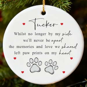 May include: A white ceramic Christmas ornament with a gold string, featuring the name "Tucker" and a heartfelt message about cherished memories and paw prints. Red hearts and paw prints decorate the design.
