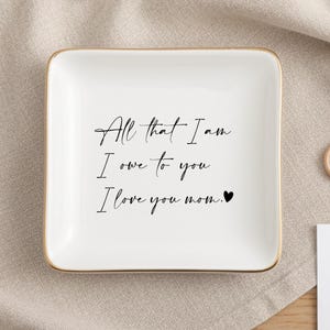 以下が含まれることがあります： 金色の縁取りが付いた白い正方形の小物入れ。 「All that I am I owe to you I love you mom」と書かれています。 金色の指輪と「Mr. &」と書かれたカードも写っています。