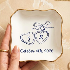Può includere: Un vassoio portagioie in ceramica bianca con bordo dorato, con due medaglioni a forma di cuore con le lettere "J" ed "E" e la data "4 ottobre 2026" in blu navy. Sono visibili anche un anello e una collana d'oro.
