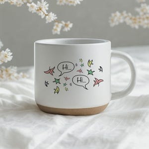 Pode incluir: Uma caneca de cerâmica branca com acabamento salpicado e base na cor de argila natural. A frente apresenta um gráfico de folhas coloridas e estilizadas espalhadas em torno de dois balões de fala, cada um contendo o texto preto "Hi..." em fonte simples.