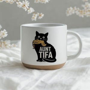 Può includere: Una tazza in ceramica bianca con finitura maculata e base color cuoio. Il lato anteriore presenta una grafica nera di un gatto che tiene in bocca un paio di baffi color cuoio, con il testo AUNT TIFA stampato in lettere maiuscole nere.