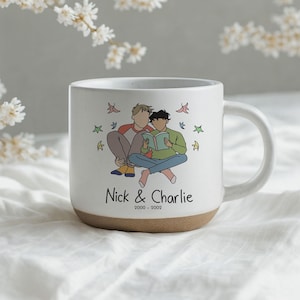 Pode incluir: Uma caneca de cerâmica branca com acabamento salpicado e base bege. A frente apresenta uma ilustração de linhas de duas pessoas sentadas lendo um livro, rodeadas por pequenas folhas coloridas. O texto diz: Nick & Charlie 2000 - 2002.