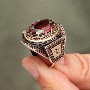 May include: A silver ring with a large, oval-cut, red gemstone. The ring features a gold-colored setting around the stone and smaller red stones. A gold-colored rectangular plate with the letter "M" is on the side.