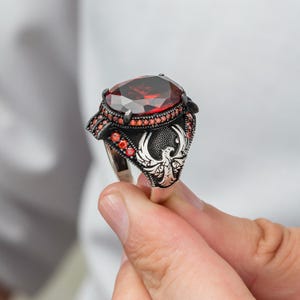 Op de afbeelding: Een zilveren ring met een grote, ovaal geslepen, rode edelsteen. De ring heeft een gedetailleerd ontwerp met een feniksembleem op de voorkant en kleinere rode stenen rond de hoofdsteen. De ring wordt door een hand omhoog gehouden.