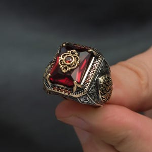 Peut inclure: Une bague en argent ornée d'une grande pierre rouge foncé rectangulaire. La bague présente des filigranes complexes et des accents dorés autour de la pierre et sur l'anneau, créant une esthétique vintage ou antique.
