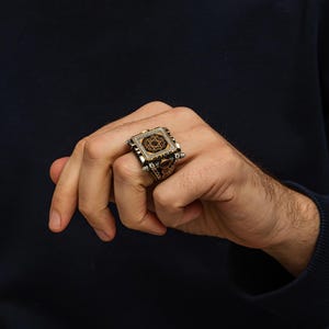 May include: A silver ring with a square face featuring a gold-colored Star of David design. The ring has intricate detailing and is set with small, reddish-brown stones. The ring is worn on a hand with a dark blue sleeve in the background.