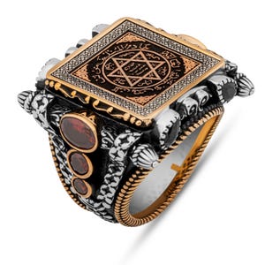 May include: A detailed silver ring with gold accents and a central square design. The square features a star symbol and intricate patterns. Three red gemstones are set on the side. The ring has a textured band and ornate detailing.