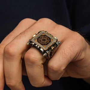 May include: A detailed silver ring with a square face featuring a six-pointed star design within a diamond shape. The ring has intricate side detailing and a gold-colored border around the central design. The ring is worn on a finger.