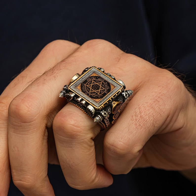 May include: A detailed silver ring with a square face featuring a six-pointed star design within a diamond shape. The ring has intricate side detailing and a gold-colored border around the central design. The ring is worn on a finger.