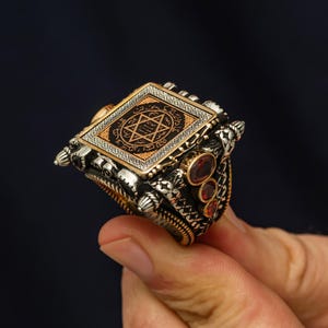 May include: A silver ring with a square, gold-toned centerpiece featuring a six-pointed star design and intricate script. The ring is adorned with three red gemstones and detailed metalwork.