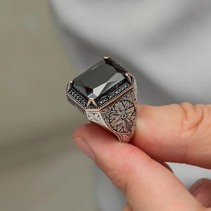 May include: A silver ring with a large, rectangular black gemstone. The ring features intricate floral engravings on the band and is accented with smaller black stones around the main gem. The setting has gold-colored accents.