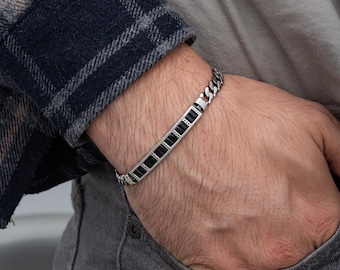 May include: A silver bracelet with a chain link design and a rectangular black stone accent. The bracelet is worn on a wrist, with a plaid shirt sleeve visible in the background.