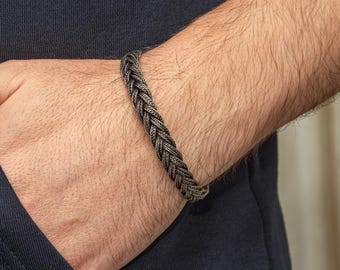 May include: Braided silver bracelet. The bracelet is made of interwoven strands of silver-toned metal, creating a textured, rope-like appearance. The bracelet is worn on a person's wrist, against a dark blue shirt.