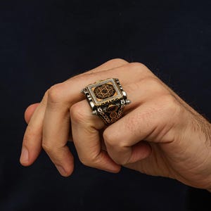 May include: A silver ring with a rectangular face featuring a gold-colored design of a star within a circle, surrounded by small white stones. The ring has intricate side detailing and a reddish-brown stone accent.