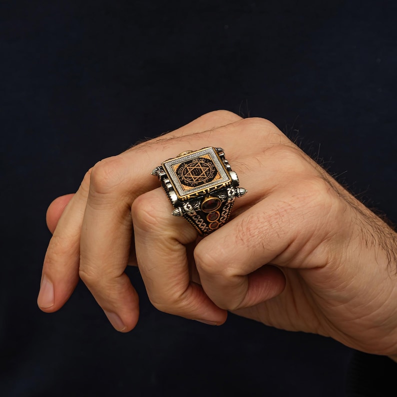 May include: A silver ring with a rectangular face featuring a gold-colored design of a star within a circle, surrounded by small white stones. The ring has intricate side detailing and a reddish-brown stone accent.
