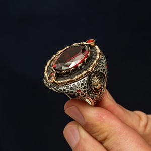 May include: A silver and gold-toned ring featuring a large oval-cut dark red gemstone in the center. The band is decorated with intricate metalwork, including a lion head relief, small red accent stones, and geometric star patterns.