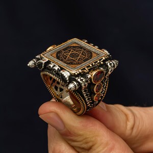 May include: A detailed silver ring with a square top featuring a geometric design and a star symbol. The ring has gold accents and three red gemstones on the side. The ring is held by a hand.