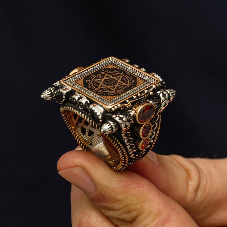 May include: A detailed silver ring with a square top featuring a geometric design and a star symbol. The ring has gold accents and three red gemstones on the side. The ring is held by a hand.