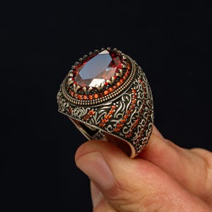 May include: A silver ring with an oval, deep red gemstone. The ring features intricate filigree and small red accent stones around the main stone and on the band. The ring is held against a dark background.