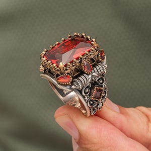 Op de afbeelding: Zilveren ring met een grote, rechthoekige, rode edelsteen. De ring heeft gedetailleerde goudkleurige filigraan en kleinere rode stenen. Het ontwerp is sierlijk en gedetailleerd, met een vintage of antieke uitstraling.