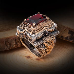 Può includere: Un anello d'argento dettagliato con accenti dorati e una grande pietra preziosa rettangolare rosso scuro. L'anello presenta intricati filigrane ed elementi decorativi, che mostrano uno stile vintage o antico. La gemma misura circa 1,3 cm di lunghezza.